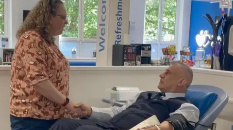 A man and woman shaking hands. The man is lying down a blood donation bed. He is donating plasma so is attached to a machine. The woman is standing next to the bed. She is wearing an orange top and has curly hair.
