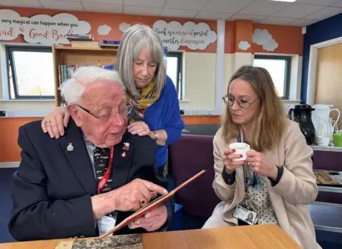 A man with grey hair, glasses and a blue jacket reading from a notepad to two women who are stood and sat next to him.