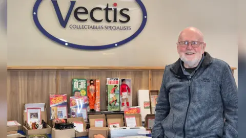 Bob Brechin, a bald man with a white beard wearing a grey zipped jacket, stands in front of a display of toys including Action Man and GI Joe figures