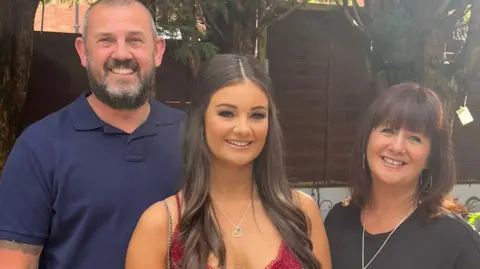 Family handout A family of three. All are smiling. A man with short, dark grey hair and a beard is on the left. In the middle is a young woman with long brown curled hair. On the right is a woman with brown short hair and a fringe.