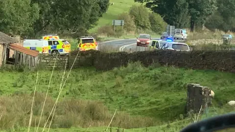 A number of police vehicles are parked next to a shed and wooden gate on the bend of a road with a field in the foreground 