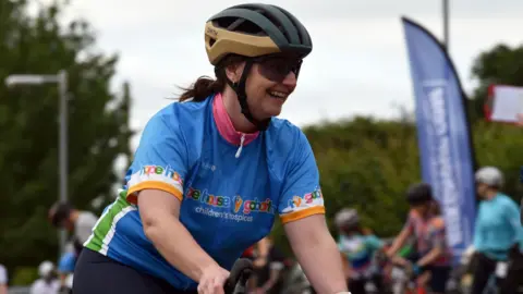 A woman with brown hair in a ponytail is smiling as she rides a bike. She is wearing black cycling glasses, a black and gold helmet and a blue t-shirt that reads "Hope House children's hospices"