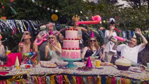 A wide shot of a long table with people gathered around celebrating. They are wearing party hats and playing with balloons. There is also a three-tier cake in the middle of the table.