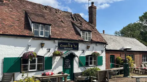 A view of the Crooked Billet Stoke Row on a sunny day. The damage to the roof following the fire is visible. The area around the building is taped.