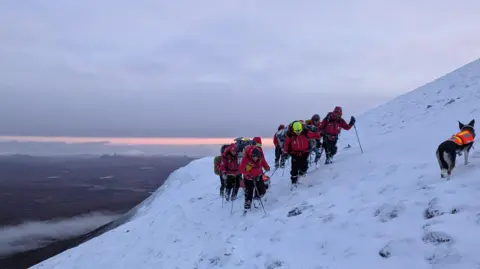Assynt Mountain Rescue A group of people in red coats and mountain gear are walking on the slope of a mountain that is covered in white snow. There is a dog in an orange vest with them. They are dragging a man in a red stretched and have walking poles. In the distance you can see land all around and clouds below.