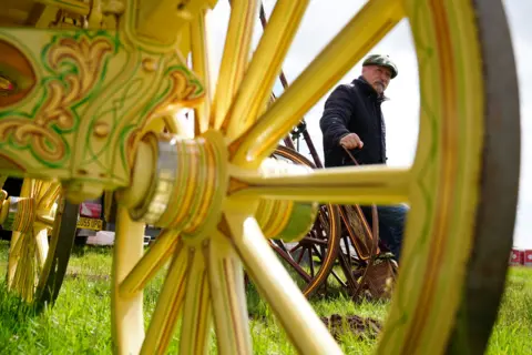 Owen Humphreys/PA Media Billy Welch pictured through the yellow spokes of a carriage wheel. He is wearing a cap and a dark coat. He has a grey beard.