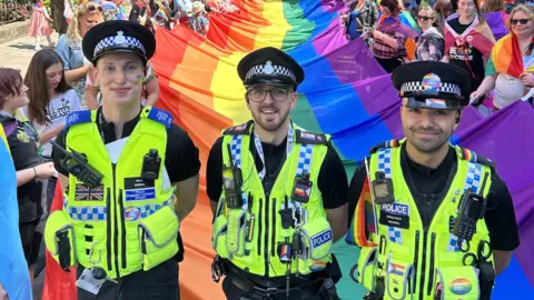 North Yorkshire Police Three police officers stand smiling in front of a large pride flag held by members of the public at York Pride 2023. They are in uniform, with pride flag stickers and patches adorning their vests. One officer has a pride flag painted onto her cheek.