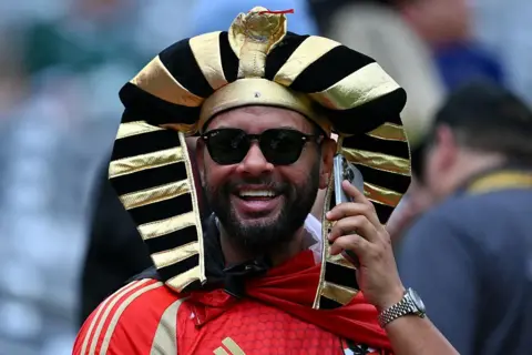 ANGELA WEISS / AFP / GETTY IMAGES A man wearing a fancy dress, pharaoh-style hat smiles and talks on the phone during the match.