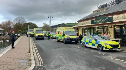 Multiple police and ambulance vehicles are parked on the road near the river. The Embankment Bistro on the right hand side. A man, wearing black, is stood watching the emergency services.