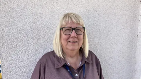 Linda has shoulder length blonde hair and is waring glasses. She is standing by a white wall which is behind her and is wearing a lanyard around her neck.