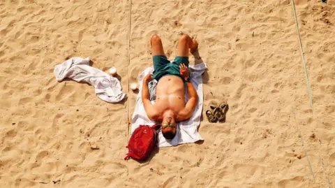 A man sunbathes on a sandy beach. The man is wearing green swimming shorts and sunglasses and he is laying on a towel. There is another towel beside him as well as a small red rucksack and a pair of brown sandals.