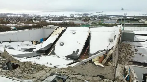 RTÉ The roof of a large building has collapsed inwards with bricks spilled out on the sides. there is snow on the roof and rubble can be seen surrounding the building. 