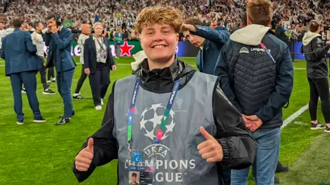 Abby Morton is wearing a grey bib, black jacket and lanyard and is on a football pitch with her thumbs up. She has short brown hair and is smiling. Behind her in the stands are hundreds of fans.