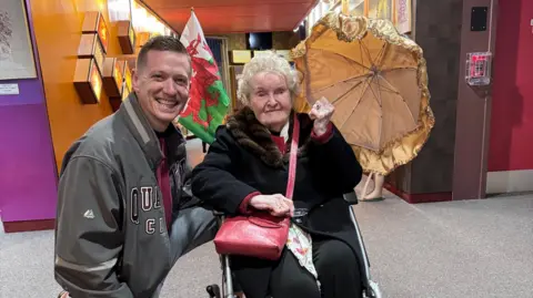 Kory Aversa Photograph of Avril Davidge (right) sitting in her wheelchair. She wears a black long coat with fur trim on the collar. She wears a red cross body bag and holds a golden umbrella in her right hand. Jimmy Good (left) wears a baseball style jacket and crouches down next to Avril. Her Welsh flag can be seen on the left hand side of her wheelchair. They both smile. 