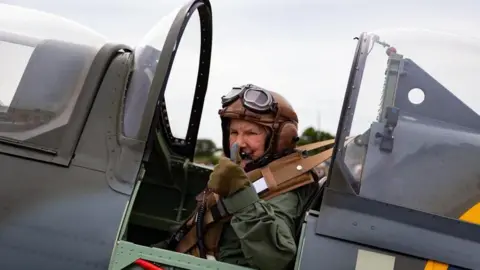 Doreen Allen, wearing a olive green flight suit, gives the thumbs up in a grey  Spitfire plane