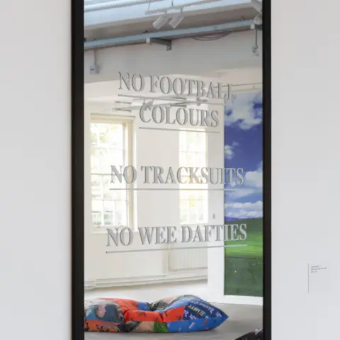 Trackie McLeod A mirror has lettering on it reading "no football colours, no tracksuits, no wee dafties". In the mirror you can see a reflection if his wider exhibition. 