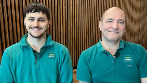 BBC Sport Wayne Meakin and his son Cole Fell-Meakin, sitting and smiling and wearing green shirts. They are in an office environment with a wooden background.