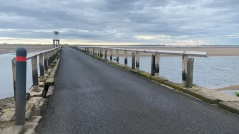 A narrow straight road bridge sits above a body of water on a tidal causeway stretching towards Holy Island which can be seen in the distance. There are straight wooden barriers, one on each side of the road, which are made of uprights at equal distances and a horizontal top bar created with long, square-ended wooden poles (like a fence posts) turned so one corner of the square cut sits on the uprights. A large expanse of light sand can be seen either side of the bridge at the far end. Halfway along the roadway is a white building which stands high above the bridge. It looks like a white garden shed on stilts and is the refuge point where people trapped by the tide can remain in safety.
