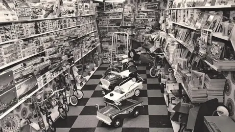 Roger Salter A black and white photo showing the inside of a toy shop with several bikes, puzzles, kits and toy cars