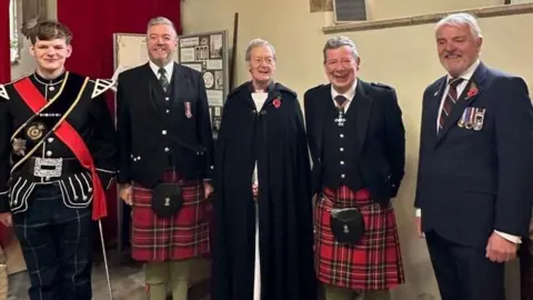David Kerr A group of pipers dressed in their kilts and tartan stand next to a vicar and another man in a suit with war medals in a church.