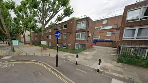 Google A Street View image showing Beachcroft Way, on the corner with Courtauld Road, with a small lamppost and two bollards in front of small blocks of flats