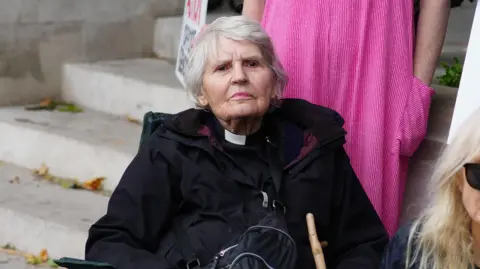 PA/ Jeff Moore Reverend Sue Parfitt sitting in a chair wearing a black raincoat and priest dog collar. She has short white hair and is looking into the distance with a stern and determined look on her face.