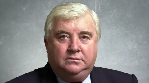 An image of Peter Kilfoyle during his time as the MP for Liverpool Walton. It is a head-and-shoulders publicity shot. He has short grey hair and is wearing a white shirt, red and black checkered tie and a navy blue suit jacket.