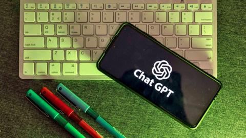 Computer keyboard with phone showing the ChatGPT logo and three coloured pens