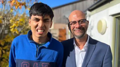 BBC Two people standing outdoors in bright sunlight, one wearing a blue GAP sweatshirt and the other in a dark blazer with a white shirt, with autumn trees and a building in the background.