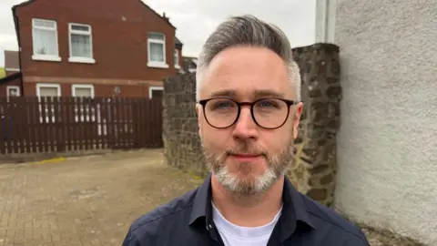 Gary has grey hair, lifted into a high fridge. He has short grey stubble and round dark-rimmed glasses. 