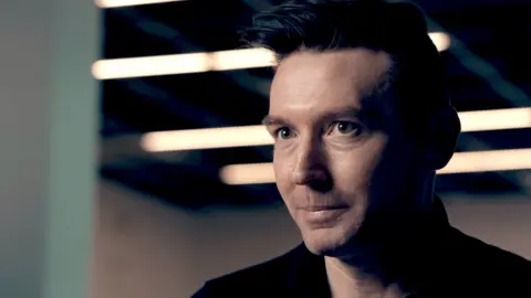 Matt Motyl, a man with brown hair styled in a quiff, pictured in close-up wearing a black shirt as he sits in an industrial-looking space with overhead strip lighting visible in the background blur.