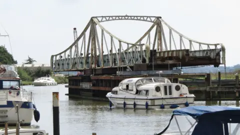 Colin Smith/Geograph Reedham Swing Bridge open for river traffic. Blue and white boats are moored on the riverbank and a motor cruiser passes along the river