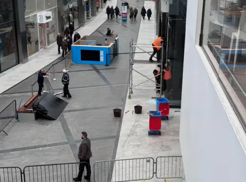 Workers in high‑visibility clothing securing damaged glass doors with scaffolding inside the Almondvale Shopping Centre in Livingston, with barriers and debris on the ground after a ram‑raid incident.