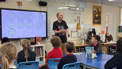 Chris Walker stands in front of a whiteboard speaking to a classroom of children aged about nine.