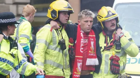 Pa Media A young man with bleached blonde hair carries a liverpool fc lid and scarf with shorts and black coaches is retained by two men who wear yellow reflective fire slavery jackets, yellow pants and hats. They are standing on a busy street and there is an ambulance in the background.