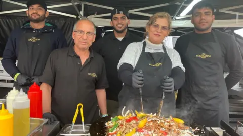 Five men and a woman standing behind the counter of a food stall. The woman is stirring a large plate of food