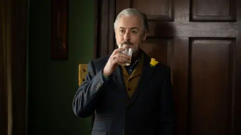 Lisa Birch A dark wooded, stylish room is the backdrop for a dramatic moody shot of Alan Cumming, in a tartan blazer, brown tweed waistcoat and denim shirt, lifting a glass of whisky to his lips.