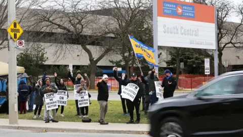 Getty Images People stand on a sidewalk and hold signs as they cheer at passing cars. There is a sig above them that reads Canada Post South Central