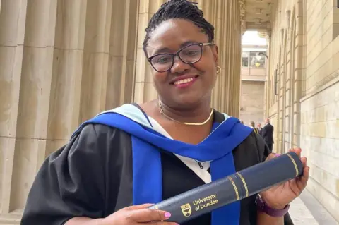 Police Scotland Dr Gomo, who is wearing black graduation robes, smiles as she holds a University of Dundee scroll. She is also wearing glasses.