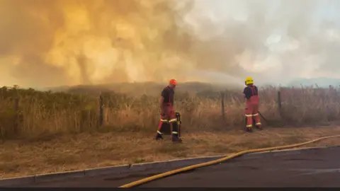 Two firefighters, one using a hose to spray water over a burning field. A haze of deep yellow smoke hangs over an area of dry, brown grass.The two men are wearing red, high-vis overalls, one wears a yellow protective helmet, and the other wears a red baseball cap.