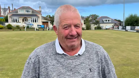 Richard Jones is stood in front of a large grassy field. He has short grey hair and is smiling and looking at the camera. 