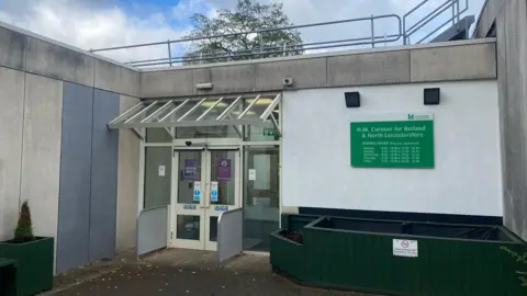 A photograph of glass double doors, with white frames to Rutland and North Leicestershire coroner's court. Next to the doors, on the wall is a green sign saying "H.M Coroner for Rutland and North Leicestershire". 