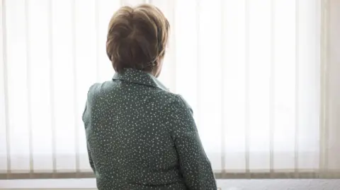Stock image of a woman with short hair wearing a grey/green dressing gown with white spots on it, looking out towards a window covered in vertical blinds