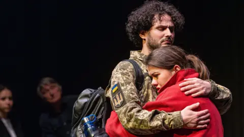 York St John University/Supplied A male actor with curly hair and a beard, dressed in military uniform, embraces a brown-haired female actor wearing a red jumper. In the background, two audience members can be seen watching.