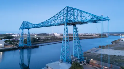 Historic England The blue Transporter Bridge, sits across a river at dusk. Buildings can be seen either side of the river.