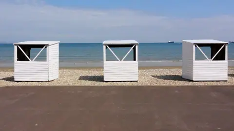 Three evenly spaced wooden beach chalets on the beach as viewed from the promenade, looking out to sea. The white painted structures have an open window at the back and no doors at the front so you can see through them. They are empty and are positioned about two or three metres apart on the shingle beach. It's a sunny day, the sea is calm and there is a ship on the horizon.