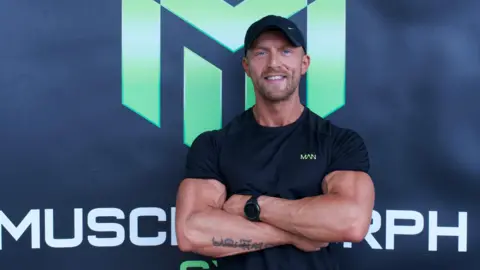 Sion Monty Sion standing with his arms crossed in front of a black branded gym backdrop. He wears a black t-shirt and a black cap. He smiles while looking at the camera. 