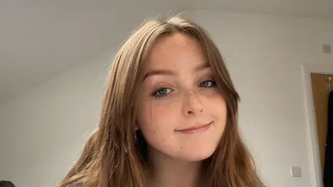 Jamie Waller A closeup of a girl with brown hair smiling with no teeth. 