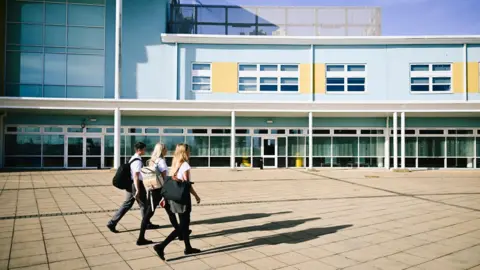 The image shows three students walking past a blue and yellow school building. They are all wearing black and white uniform.