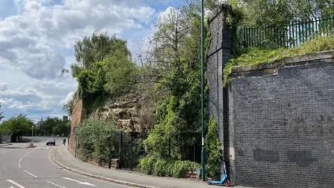 BBC The caves, which are by a main road, bordered by Victorian retaining walls and covered in bushes, as they appeared in June.